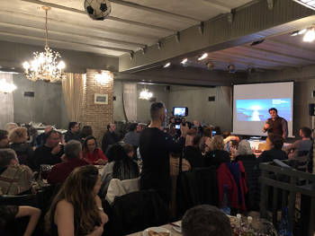 A crowd of people are sitting at tables inside a restaurant, a man is standing on the stage and talking and the photographer is filming him